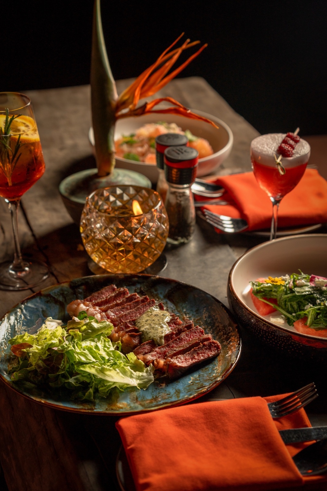 A candlelit table spread at BABOU, sliced steak, salad, and cocktails